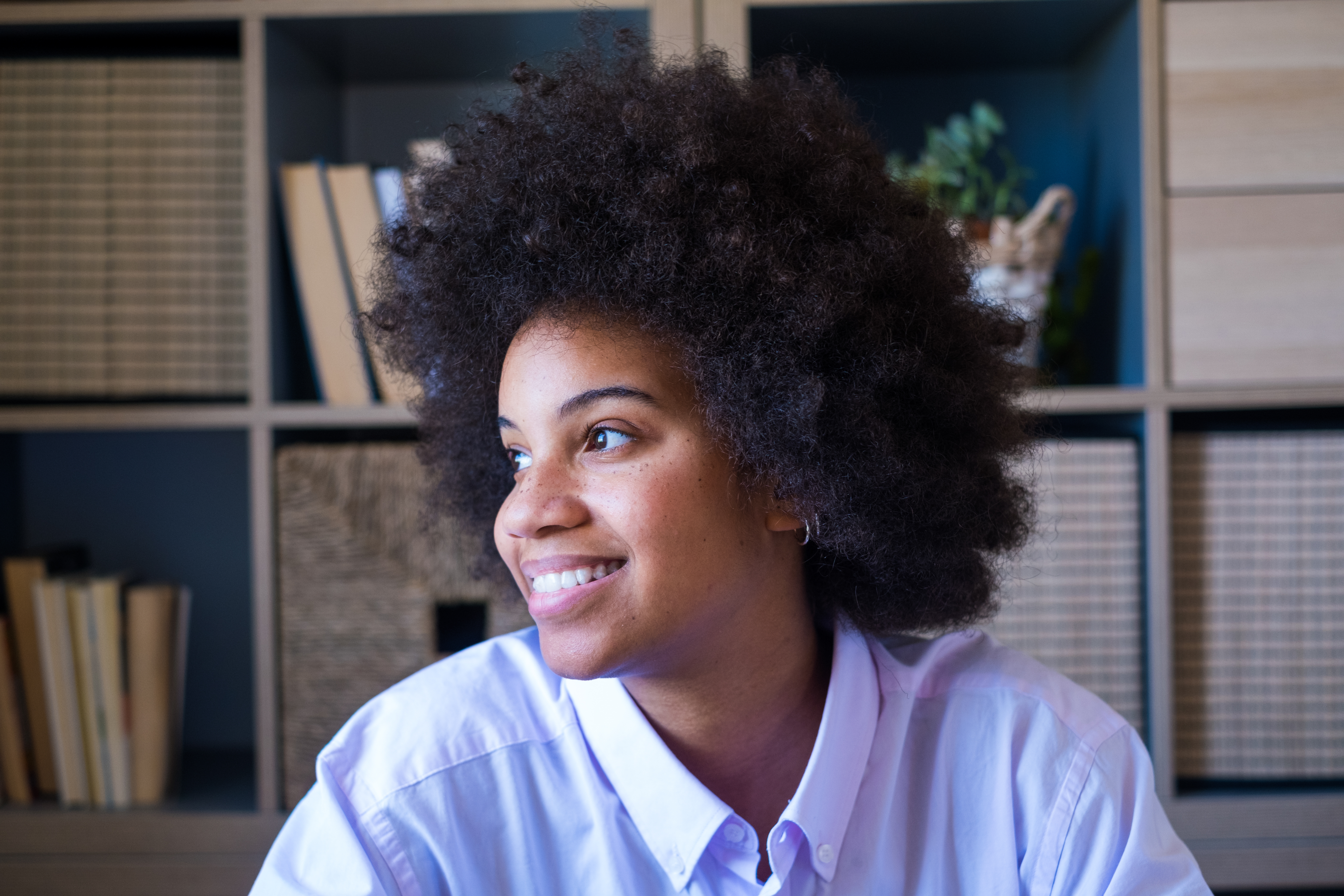 Attractive dark skinned young woman with curly Afro hairstyle looking out through window. Thoughtful african american businesswoman smiling and looking away at office. Female lady dreaming about future