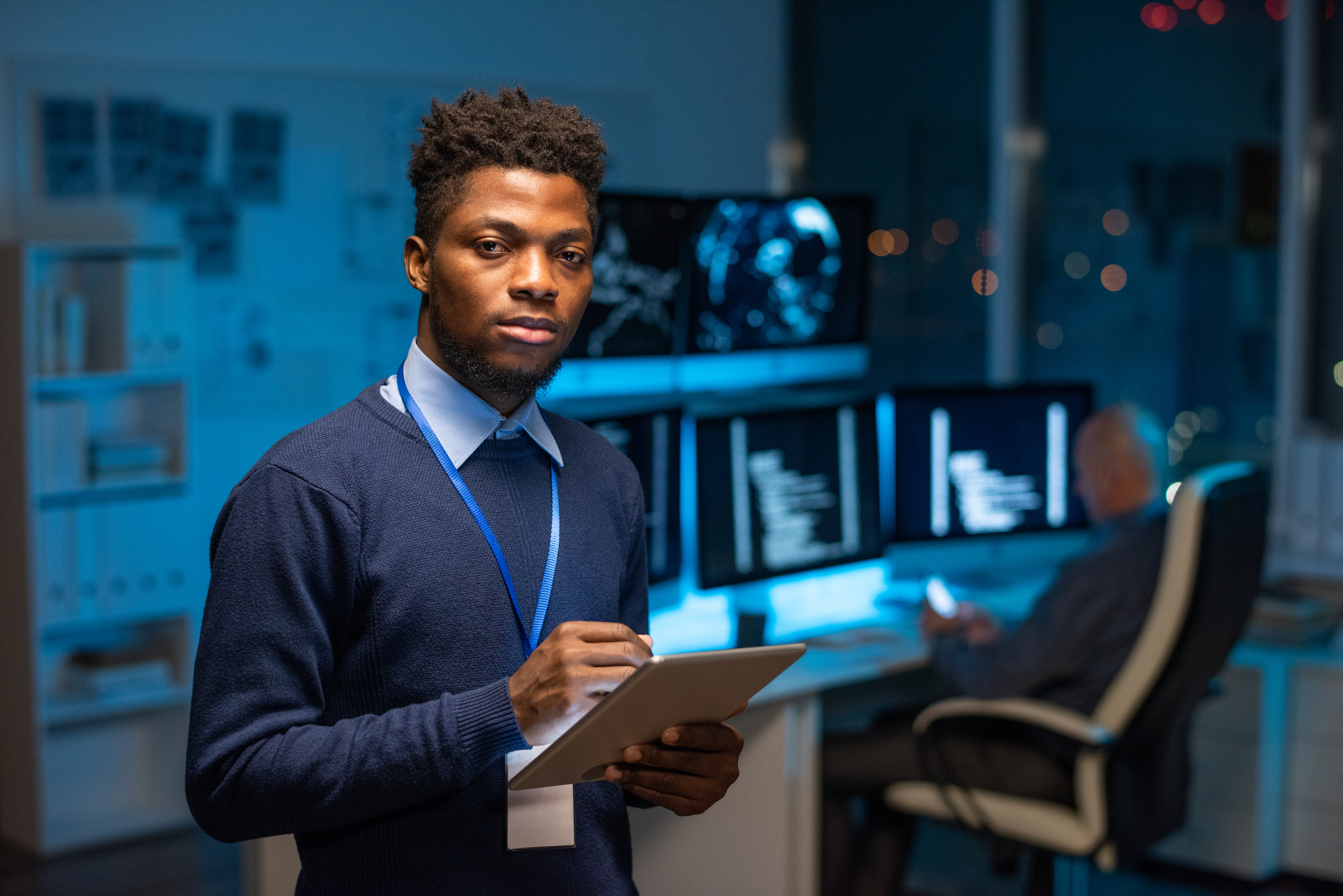Young serious African programmer with tablet standing in front of camera against colleague by workplace